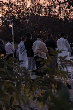 A group of people standing in an outdoor setting during sunset. They appear to be engaged in a communal or social activity. The background shows trees silhouetted against the evening sky, and the ground is partially obscured by bushes in the foreground.