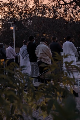 A group of people standing in an outdoor setting during sunset. They appear to be engaged in a communal or social activity. The background shows trees silhouetted against the evening sky, and the ground is partially obscured by bushes in the foreground.