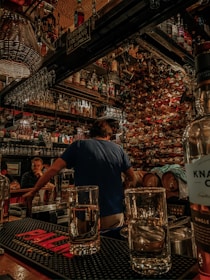 A cozy bar setting with an abundance of liquor bottles lining the walls and shelves, creating a warm and inviting atmosphere. A bartender, seen from the back, is preparing drinks at the bar counter, which is adorned with glasses and a prominent red 'PALM' mat. The ambiance is intimate, with dim lighting and wooden decor, while patrons are engaged in conversations in the background.