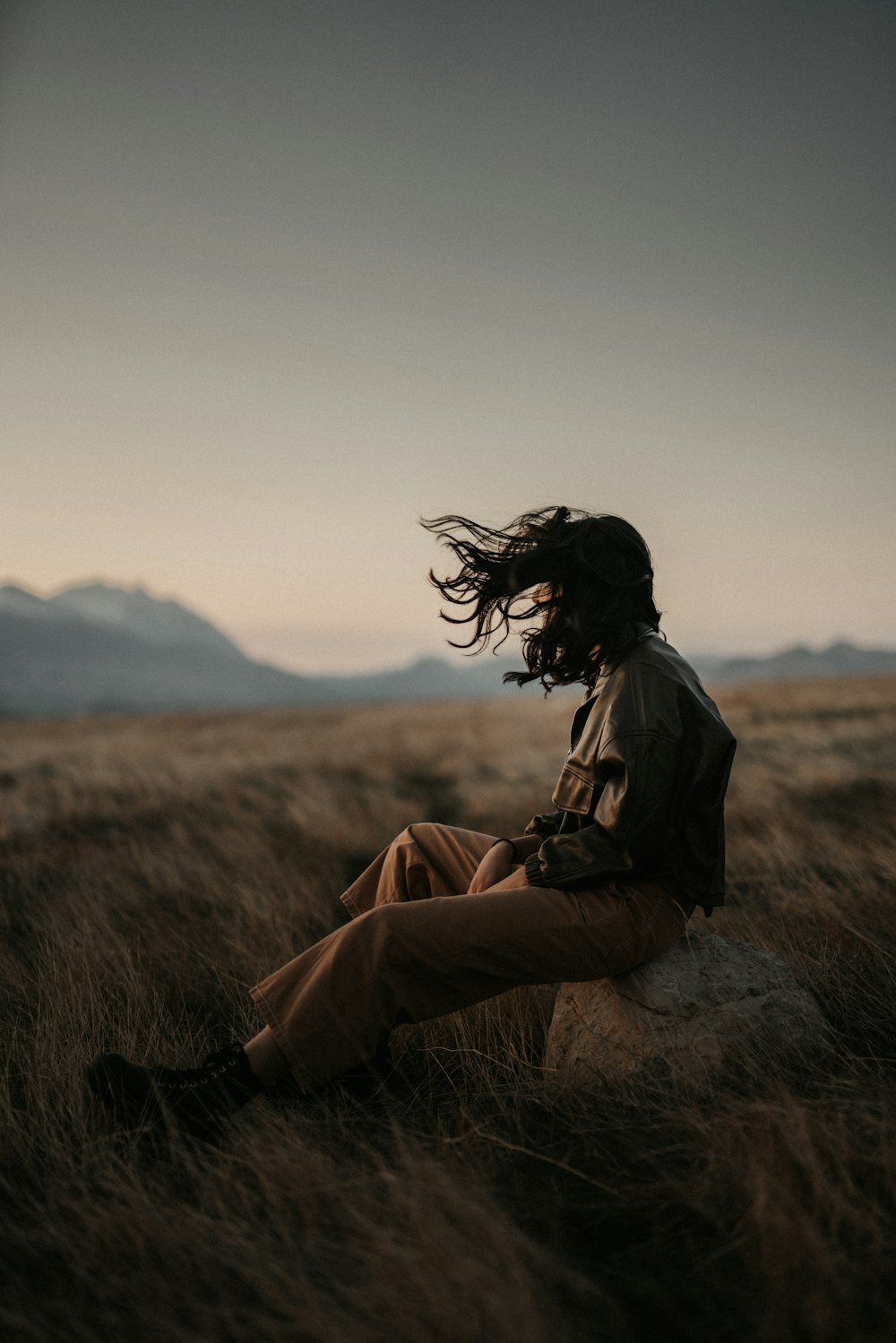 mulher na jaqueta marrom e calças marrons sentadas no campo de grama marrom durante o dia