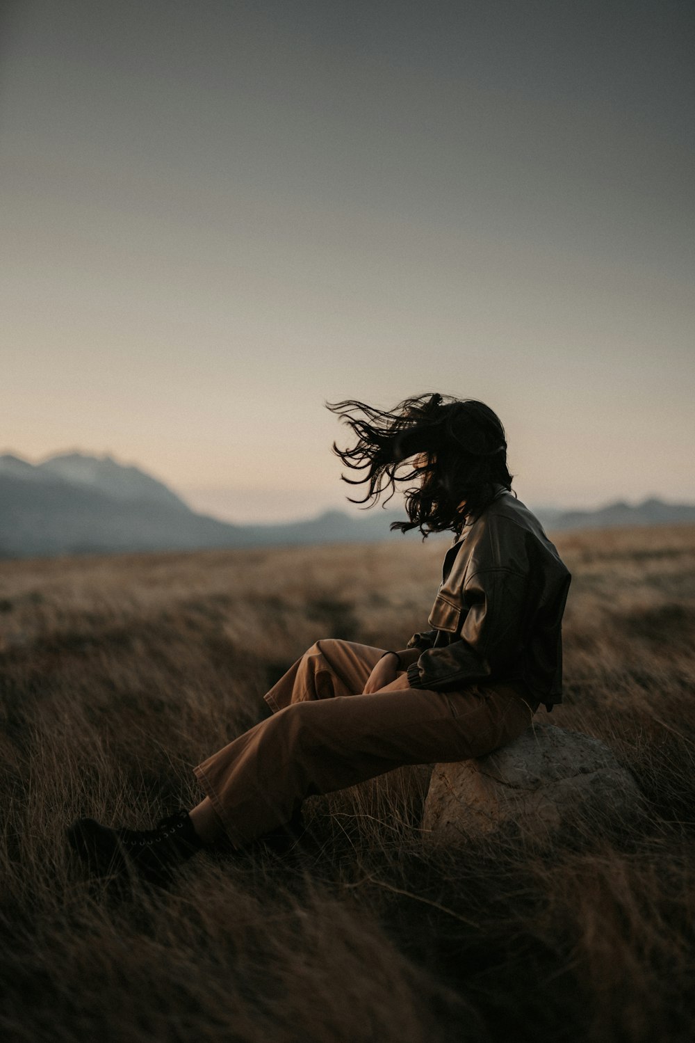 woman in brown jacket and brown pants sitting on brown grass field during daytime