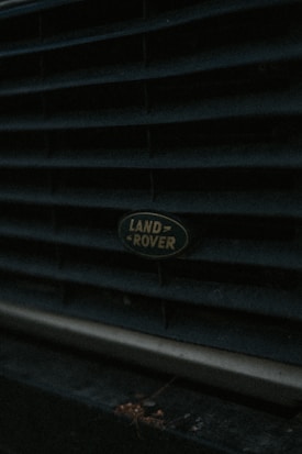 Close-up view of a Land Rover badge on the grille of a vehicle. The image is dark with subdued lighting, emphasizing the texture of the grille.