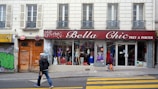A street scene featuring a clothing store named 'Bella Chic Pret a Porter' with a red awning above the window display. Mannequins with various outfits are visible through the large windows. Two pedestrians walk in front of the store, one wearing a hoodie and jeans, the other in a headscarf carrying an orange bag. The building has a classic European facade with shutters and balconies. Graffiti is present on the adjacent doors.