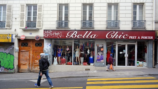 A street scene featuring a clothing store named 'Bella Chic Pret a Porter' with a red awning above the window display. Mannequins with various outfits are visible through the large windows. Two pedestrians walk in front of the store, one wearing a hoodie and jeans, the other in a headscarf carrying an orange bag. The building has a classic European facade with shutters and balconies. Graffiti is present on the adjacent doors.