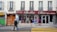A street scene featuring a clothing store named 'Bella Chic Pret a Porter' with a red awning above the window display. Mannequins with various outfits are visible through the large windows. Two pedestrians walk in front of the store, one wearing a hoodie and jeans, the other in a headscarf carrying an orange bag. The building has a classic European facade with shutters and balconies. Graffiti is present on the adjacent doors.