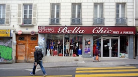 A street scene featuring a clothing store named 'Bella Chic Pret a Porter' with a red awning above the window display. Mannequins with various outfits are visible through the large windows. Two pedestrians walk in front of the store, one wearing a hoodie and jeans, the other in a headscarf carrying an orange bag. The building has a classic European facade with shutters and balconies. Graffiti is present on the adjacent doors.