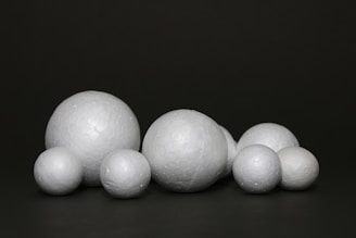 A collection of white spherical objects made of a material resembling polystyrene is arranged against a dark background. The spheres vary in size and have a textured surface.