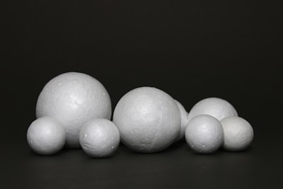 A collection of white spherical objects made of a material resembling polystyrene is arranged against a dark background. The spheres vary in size and have a textured surface.