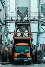 An industrial scene featuring a large orange cement truck positioned beneath a series of tall, cylindrical silos with metal framework. Workers can be seen on the upper platform surrounding the silos. The environment suggests a construction or manufacturing site with an urban, gritty atmosphere.