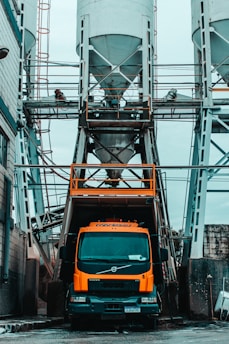 An industrial scene featuring a large orange cement truck positioned beneath a series of tall, cylindrical silos with metal framework. Workers can be seen on the upper platform surrounding the silos. The environment suggests a construction or manufacturing site with an urban, gritty atmosphere.