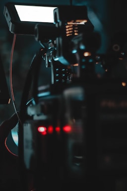 A close-up view of a complex device or machine, possibly a camera or filming equipment. The image features various controls, cables, and a monitor screen. The lighting is dim, with visible glowing red and blue lights that enhance the technological feel.
