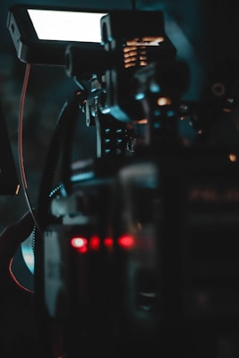 A close-up view of a complex device or machine, possibly a camera or filming equipment. The image features various controls, cables, and a monitor screen. The lighting is dim, with visible glowing red and blue lights that enhance the technological feel.