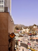 A cityscape view depicting a bustling urban area with iconic buildings and the famous Hollywood sign visible in the distance. The foreground features a modern building with palm-themed signage. The scene includes several vehicles on a winding road, surrounded by palm trees and commercial buildings adorned with billboards.