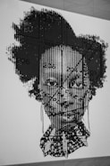 A black and white graphic artwork features a portrait of a person with a complex overlay of patterns and text. The image includes linear and textual elements, creating a textured effect on the face. The background is a plain wall, emphasizing the intricate design.