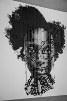 A black and white graphic artwork features a portrait of a person with a complex overlay of patterns and text. The image includes linear and textual elements, creating a textured effect on the face. The background is a plain wall, emphasizing the intricate design.