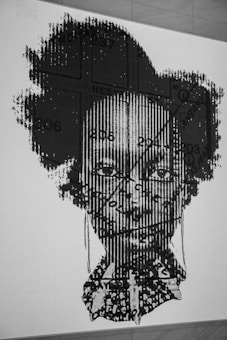 A black and white graphic artwork features a portrait of a person with a complex overlay of patterns and text. The image includes linear and textual elements, creating a textured effect on the face. The background is a plain wall, emphasizing the intricate design.