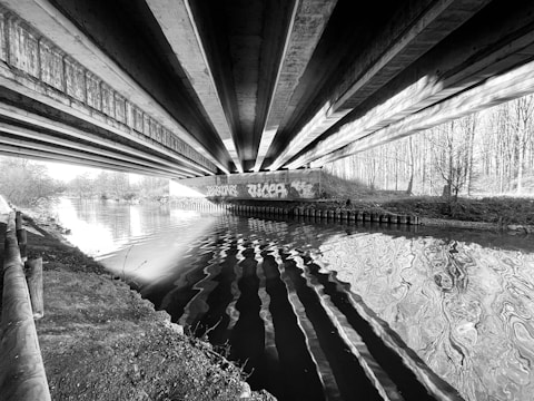 Concrete beams form an underpass above a river, with intricate reflections on the water's surface. Graffiti decorates the walls, and trees form a backdrop along the riverbank.