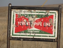 A weathered and damaged sign, mounted on a metal frame, located in a grassy field. The sign displays the words 'TORONTO PIPE LINE COMPANY' with a red and green background, overlaid with numerous bullet holes and rust marks.