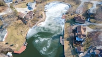 An aerial view of a small residential area featuring several houses surrounding a partially frozen lake. The lake displays areas of ice and open water, and there are docks extending into the water. The surrounding land is a mix of grassy areas and trees, with vehicles parked on driveways and roads. There are shadows indicating the time of day is either morning or afternoon.