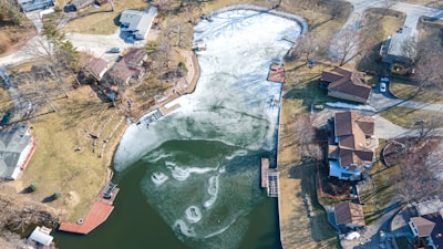 An aerial view of a small residential area featuring several houses surrounding a partially frozen lake. The lake displays areas of ice and open water, and there are docks extending into the water. The surrounding land is a mix of grassy areas and trees, with vehicles parked on driveways and roads. There are shadows indicating the time of day is either morning or afternoon.