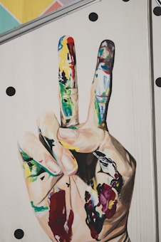 A large mural depicts a hand making a peace sign with two fingers up. The fingers and palm are covered in vibrant, multicolored paint splatters. The background is a light surface with scattered black dots.