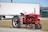 A vintage red tractor is parked on a grassy area, with a 'For Sale' sign attached to it. In the background, there is a large white trailer and a green building. The tractor is positioned on a gravel-covered path.