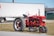 A vintage red tractor is parked on a grassy area, with a 'For Sale' sign attached to it. In the background, there is a large white trailer and a green building. The tractor is positioned on a gravel-covered path.