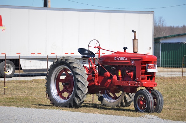 A vintage red tractor is parked on a grassy area, with a 'For Sale' sign attached to it. In the background, there is a large white trailer and a green building. The tractor is positioned on a gravel-covered path.