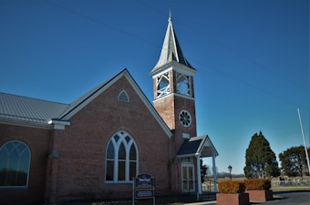 A charming brick church with a tall steeple topped by a spire, featuring arched windows and a small entrance porch. The structure is surrounded by a well-kept yard and a sign in front of the building. The sky is clear and blue, contributing to a serene atmosphere.