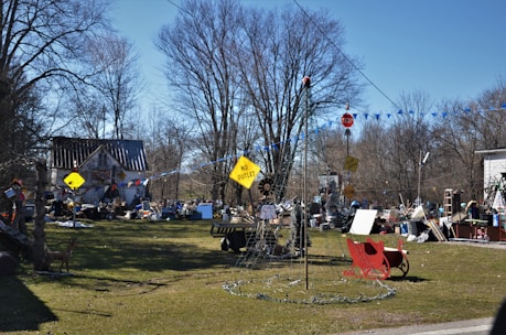 A cluttered outdoor space filled with various objects, including signs, a red sleigh, a ladder, and different piles of materials. A house with a metal roof is in the background, surrounded by leafless trees. The scene appears disorganized with numerous items scattered across the yard.