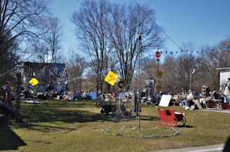 A cluttered outdoor space filled with various objects, including signs, a red sleigh, a ladder, and different piles of materials. A house with a metal roof is in the background, surrounded by leafless trees. The scene appears disorganized with numerous items scattered across the yard.