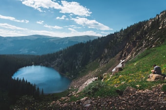 A serene mountain landscape with a clear blue lake surrounded by dense evergreen forest. The scene includes rolling hills and rocky terrain covered with patches of grass and wildflowers. A person is sitting on a rock, possibly reading or writing, adding a sense of tranquility and introspection.