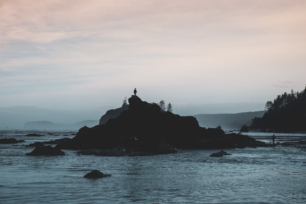 silhouette of person sitting on rock formation in the middle of sea during daytime