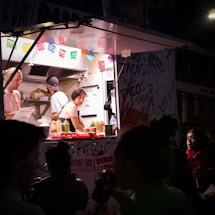 A woman working inside a food truck, preparing food in a well-lit kitchen area. Colorful papel picado banners are hanging above the counter, and bottles of condiments are neatly arranged. Outside, a group of people, some with partially visible faces, stand in the darker foreground.