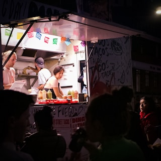 A woman working inside a food truck, preparing food in a well-lit kitchen area. Colorful papel picado banners are hanging above the counter, and bottles of condiments are neatly arranged. Outside, a group of people, some with partially visible faces, stand in the darker foreground.