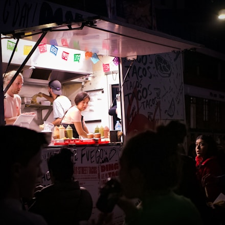 A woman working inside a food truck, preparing food in a well-lit kitchen area. Colorful papel picado banners are hanging above the counter, and bottles of condiments are neatly arranged. Outside, a group of people, some with partially visible faces, stand in the darker foreground.