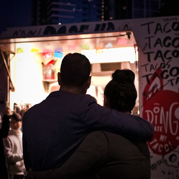A couple is standing with their backs to the camera, embracing each other in front of a brightly lit taco food truck at night. The food truck has colorful decorations and a large red circle with text around it. Other people are visible near the truck, and tall buildings can be seen in the background.