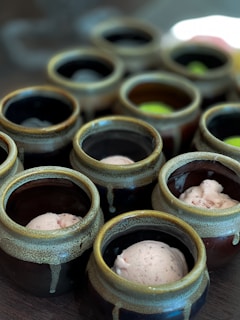 Several ceramic pots are arranged on a surface, each containing a scoop of ice cream. The pots have a rustic appearance with earthy colors and glazed textures.