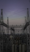 A complex network of metal towers and wires forming an electrical substation. The structure is composed of interconnected pylons and transformers, with multiple cables stretching across the frame. The sky above is a muted gradient of soft colors indicating twilight or early dawn.