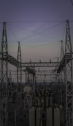 A complex network of metal towers and wires forming an electrical substation. The structure is composed of interconnected pylons and transformers, with multiple cables stretching across the frame. The sky above is a muted gradient of soft colors indicating twilight or early dawn.
