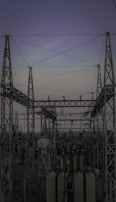 A complex network of metal towers and wires forming an electrical substation. The structure is composed of interconnected pylons and transformers, with multiple cables stretching across the frame. The sky above is a muted gradient of soft colors indicating twilight or early dawn.