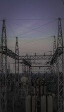A complex network of metal towers and wires forming an electrical substation. The structure is composed of interconnected pylons and transformers, with multiple cables stretching across the frame. The sky above is a muted gradient of soft colors indicating twilight or early dawn.