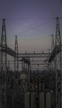 A complex network of metal towers and wires forming an electrical substation. The structure is composed of interconnected pylons and transformers, with multiple cables stretching across the frame. The sky above is a muted gradient of soft colors indicating twilight or early dawn.