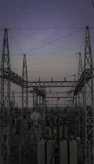 A complex network of metal towers and wires forming an electrical substation. The structure is composed of interconnected pylons and transformers, with multiple cables stretching across the frame. The sky above is a muted gradient of soft colors indicating twilight or early dawn.