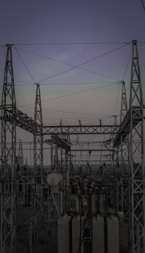 A complex network of metal towers and wires forming an electrical substation. The structure is composed of interconnected pylons and transformers, with multiple cables stretching across the frame. The sky above is a muted gradient of soft colors indicating twilight or early dawn.