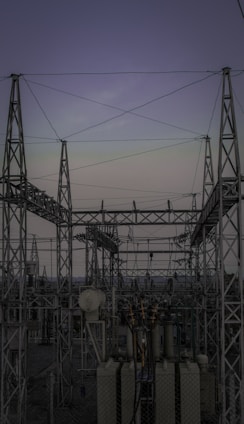 A complex network of metal towers and wires forming an electrical substation. The structure is composed of interconnected pylons and transformers, with multiple cables stretching across the frame. The sky above is a muted gradient of soft colors indicating twilight or early dawn.