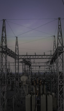 A complex network of metal towers and wires forming an electrical substation. The structure is composed of interconnected pylons and transformers, with multiple cables stretching across the frame. The sky above is a muted gradient of soft colors indicating twilight or early dawn.
