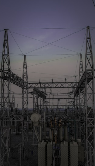 A complex network of metal towers and wires forming an electrical substation. The structure is composed of interconnected pylons and transformers, with multiple cables stretching across the frame. The sky above is a muted gradient of soft colors indicating twilight or early dawn.