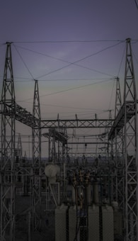 A complex network of metal towers and wires forming an electrical substation. The structure is composed of interconnected pylons and transformers, with multiple cables stretching across the frame. The sky above is a muted gradient of soft colors indicating twilight or early dawn.