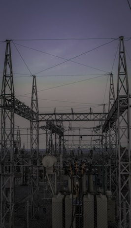 A complex network of metal towers and wires forming an electrical substation. The structure is composed of interconnected pylons and transformers, with multiple cables stretching across the frame. The sky above is a muted gradient of soft colors indicating twilight or early dawn.